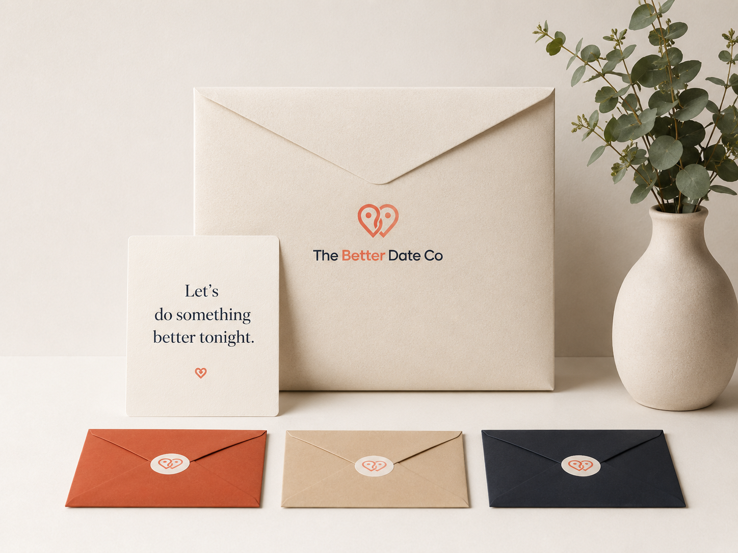The Better Date Co package: a kraft envelope printed with the logo, a note card reading 'Let's do something better tonight', and three sealed date-stage envelopes, styled next to a eucalyptus plant